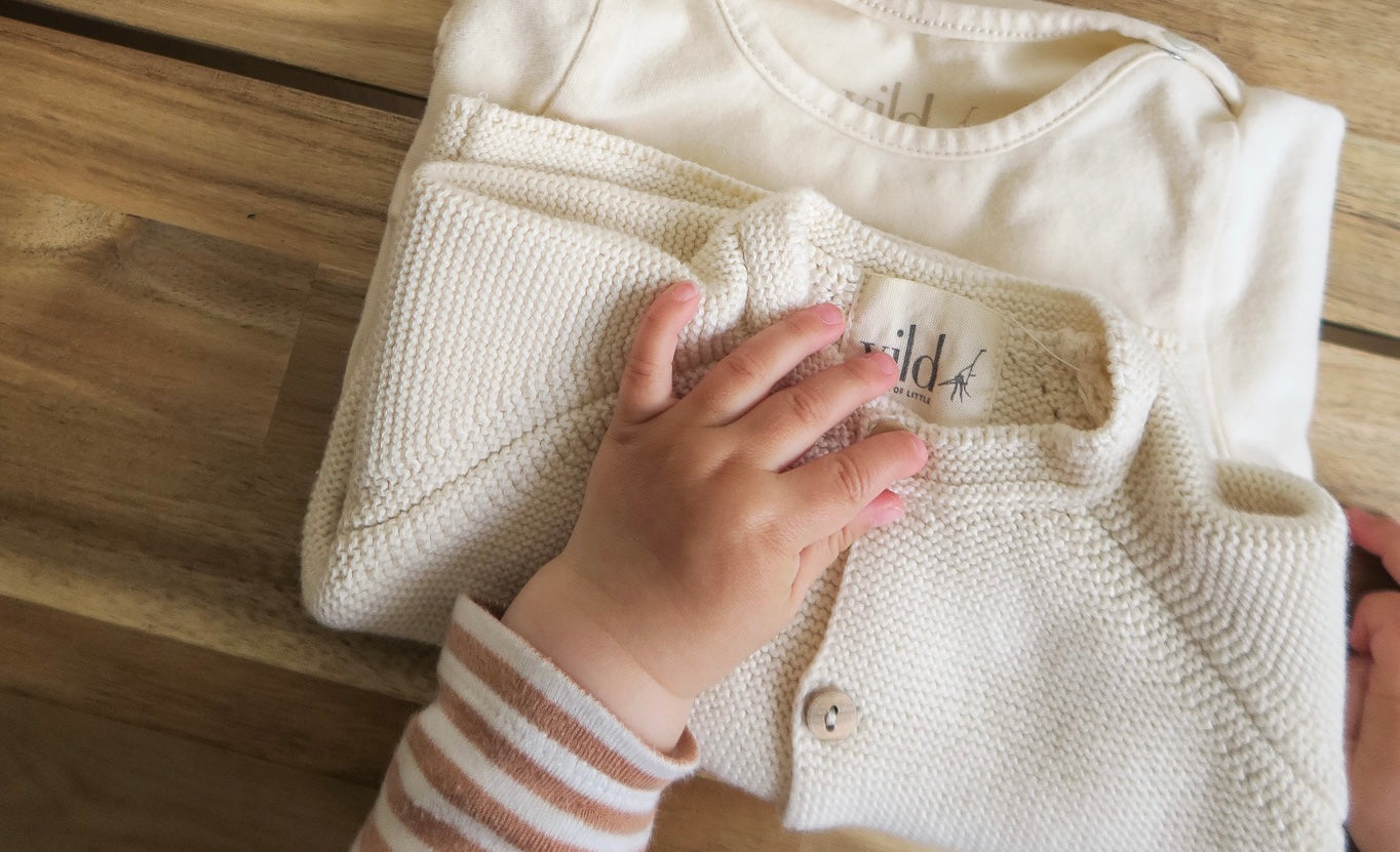 Photography from above and close-up with a wood background of an off-white knitted baby folded cardigan from OiOiOi on a off-white baby folded onesie from OiOiOi with a baby hand wearing striped brun and white long shirt from OiOiOi touching the cardigan 