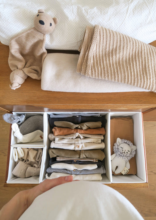 A pregnant belly with a hand on it in front of a drawer with baby clothes from OiOiOi folded in a white box with different compartment, on top of the dresser there is a brown teddy bear, a white pillow and 2 folded blankets one is white and the other is brown.