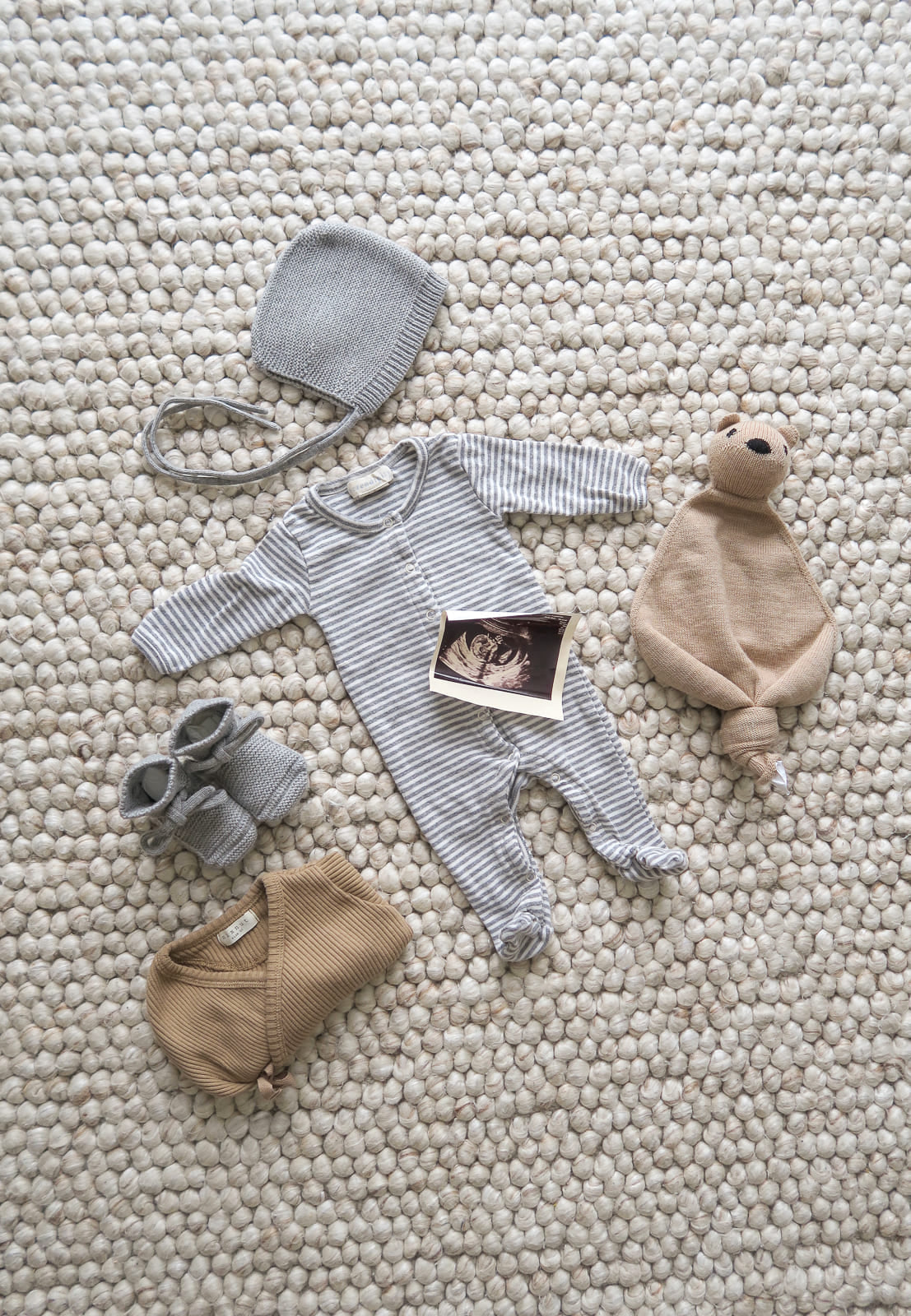 OiOiOi newborn clothes: gray cap, gray and white striped pajamas, gray shoes, brown bodysuit from OiOiOi lies on carpet