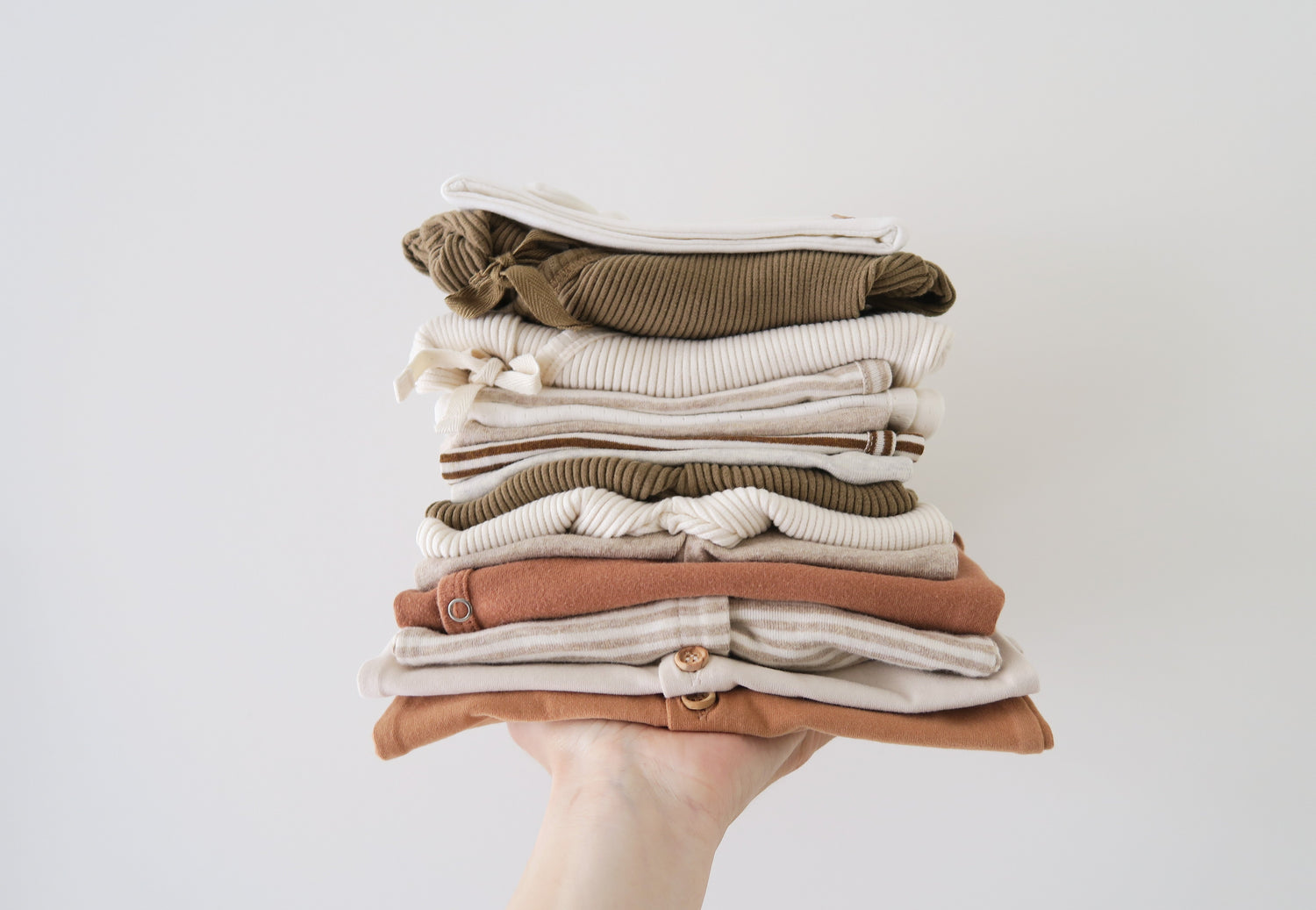 Hand with light skin color, holding folded organic cotton baby dresses in brownish and light natural colors from OiOiOi