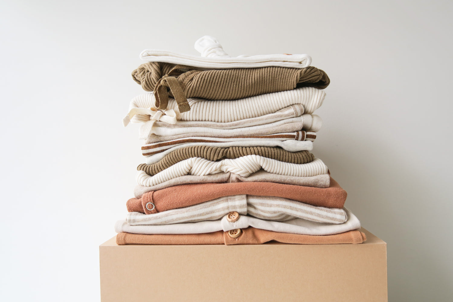 Pile of folded organic cotton baby clothes in brownish and light natural colors from OiOiOi. Bundle of baby clothes presented on a box