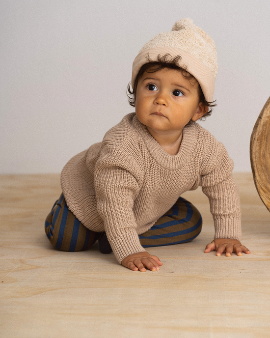 Cute baby with brown hair wearing an off-white beige hat, a brown hoodie and a stripped dark brown and blue pant from House of Jamie and OiOiOi, he is on his knees on a wood floor and white wall.