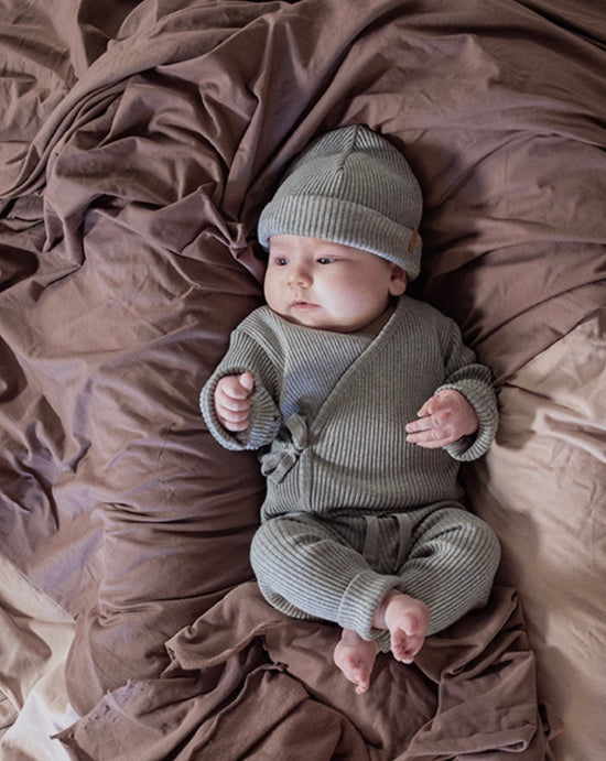 Light skinned baby with cap and clothes in khaki green from Nixnut brand and OiOiOi, lies on burgundy blanket