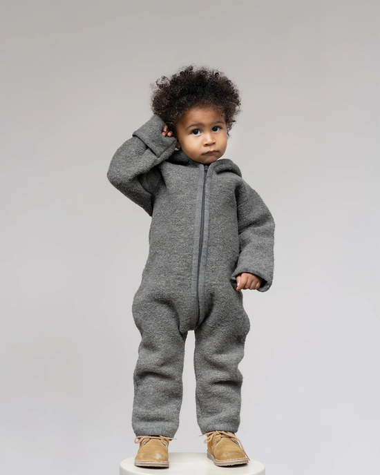 Baby with brown skin and dark curly hairs standing on a white background and wearing a grey overall from Halfen and OiOiOi. 