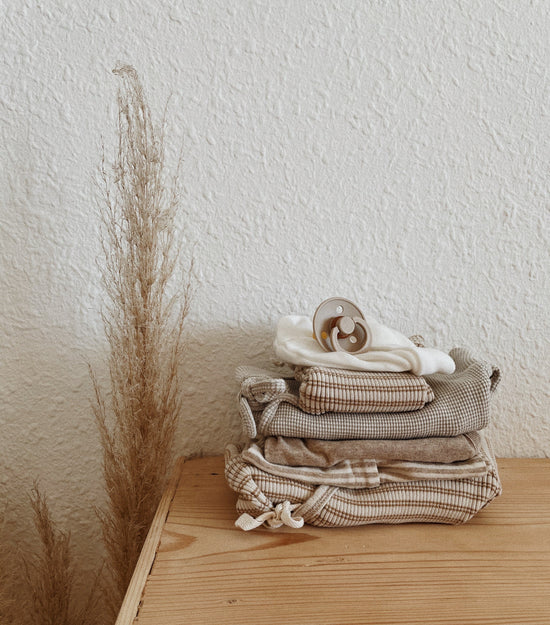 Bundle beige striped and white organic baby clothes from OiOiOi, lie on brown dresser infront of white wall