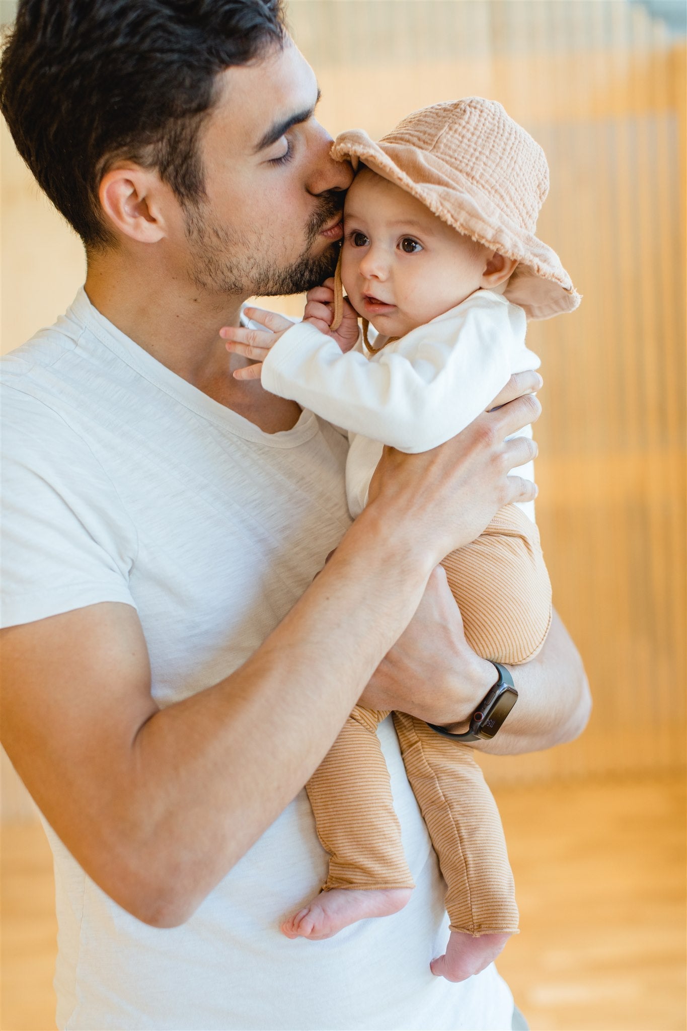 Man with white t-shirt, dark short hair and light skin color, holding baby with orange hat from OiOiOi in his arms and kissing it.